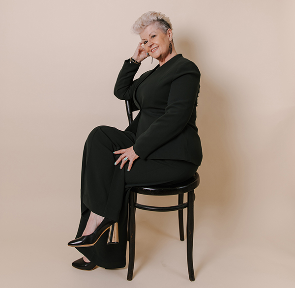Image of Chrissie wearing a green suit with black snd gold heels and sitting sideways on a black chair
